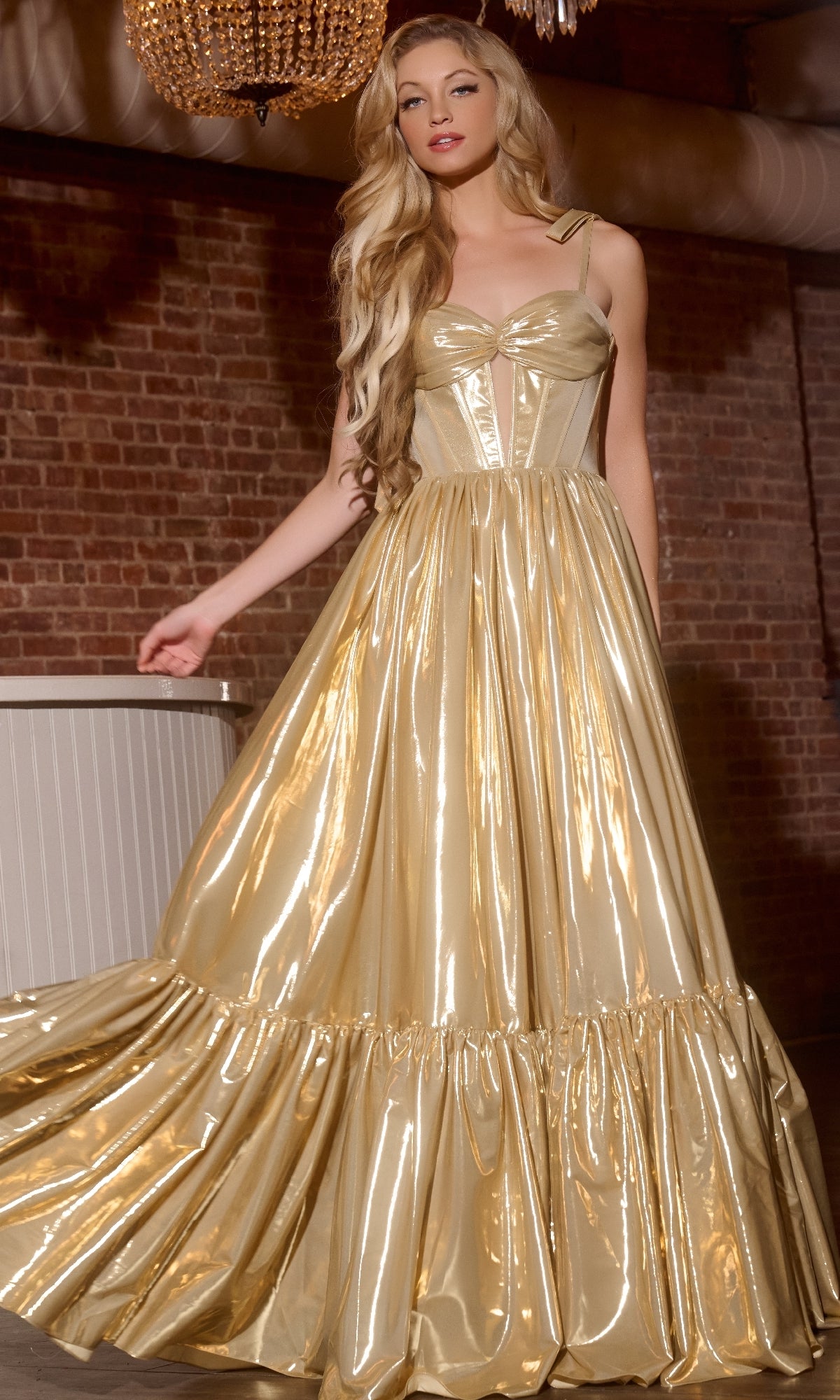 A woman poses indoors against an exposed brick wall wearing the Jovani 43596 long prom dress, featuring a shiny metallic gold finish, corset bodice, and full skirt.